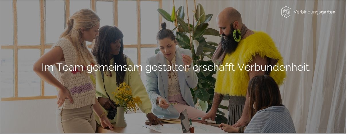 Bunt gekleidete Menschen stehen um einen Tisch herum und arbeiten kreativ. Darauf steht: Im Team gemeinsam gestalten erschafft Verbundenheit.